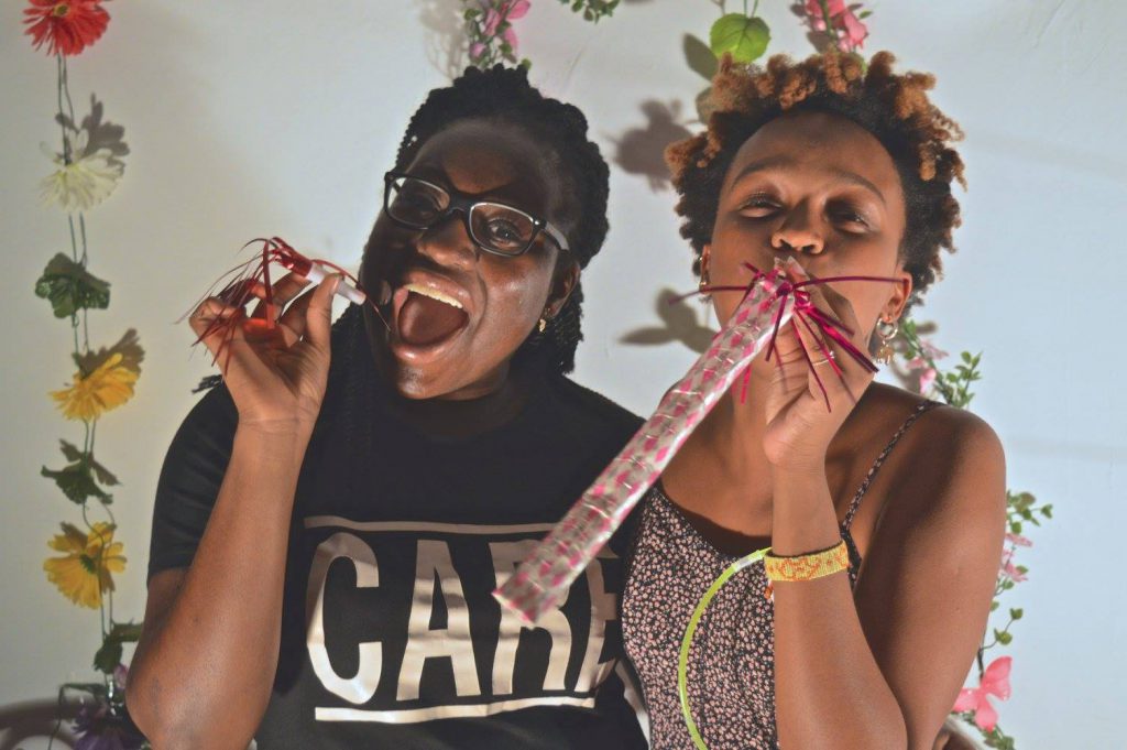 Two people blowing party horns and smiling. Behind them are four rows of flowers that are tied together.