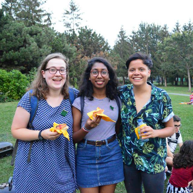 Three people holding hot dogs and smiling in a park.